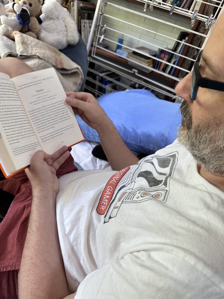 A bearded man reading a book with the chapter heading TIMPANO