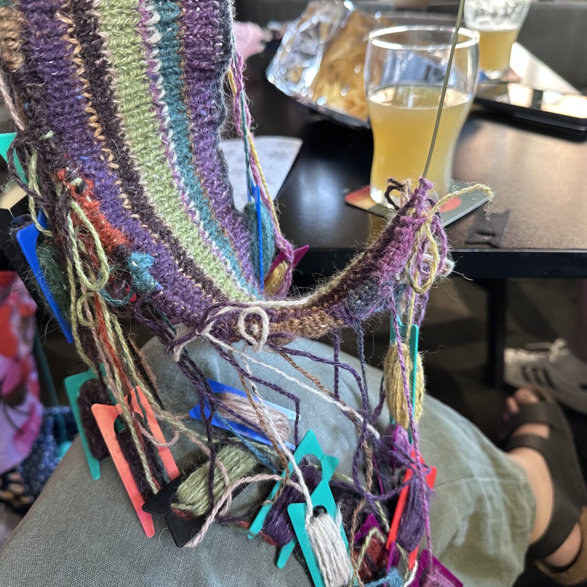 A person holding up multi-coloured knitting in a pub, with a giant tangle of dozens of coloured bobbins hanging from it