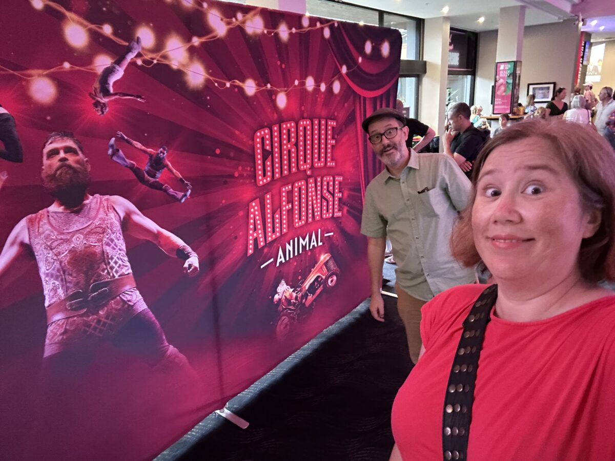 Two people in front of a large sign with circus performers on it that says CIRQUE ALFONSE - ANIMAL