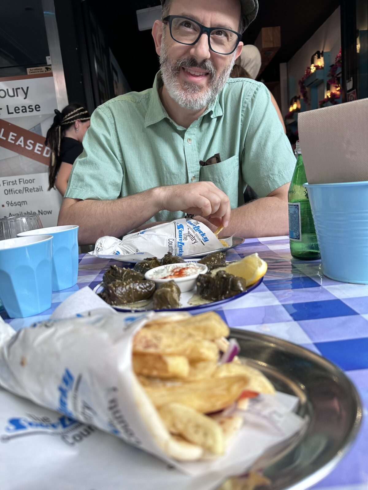 A man sits at a restaurant table with a blue and white checkered tablecloth. A plate of dolmades is in the center of the table, as well as pitas wrapped in paper.