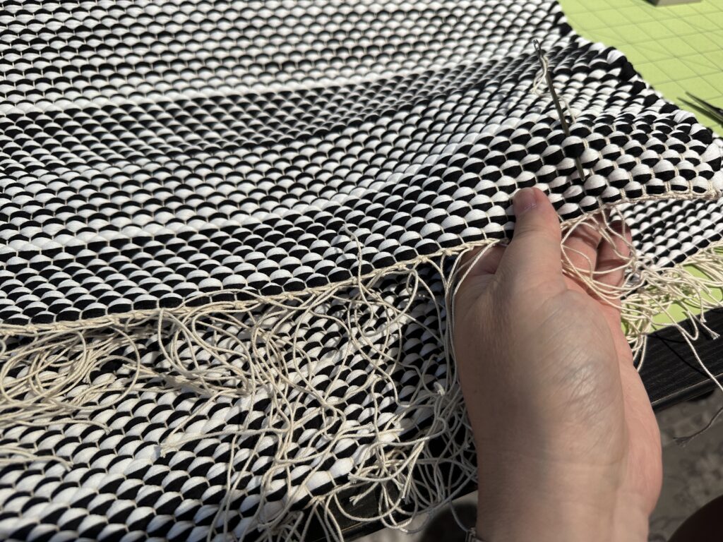 A person’s hands laboriously weaving in every warp thread from a black and white handwoven rug. There sees literally hundreds of threads to finish off.