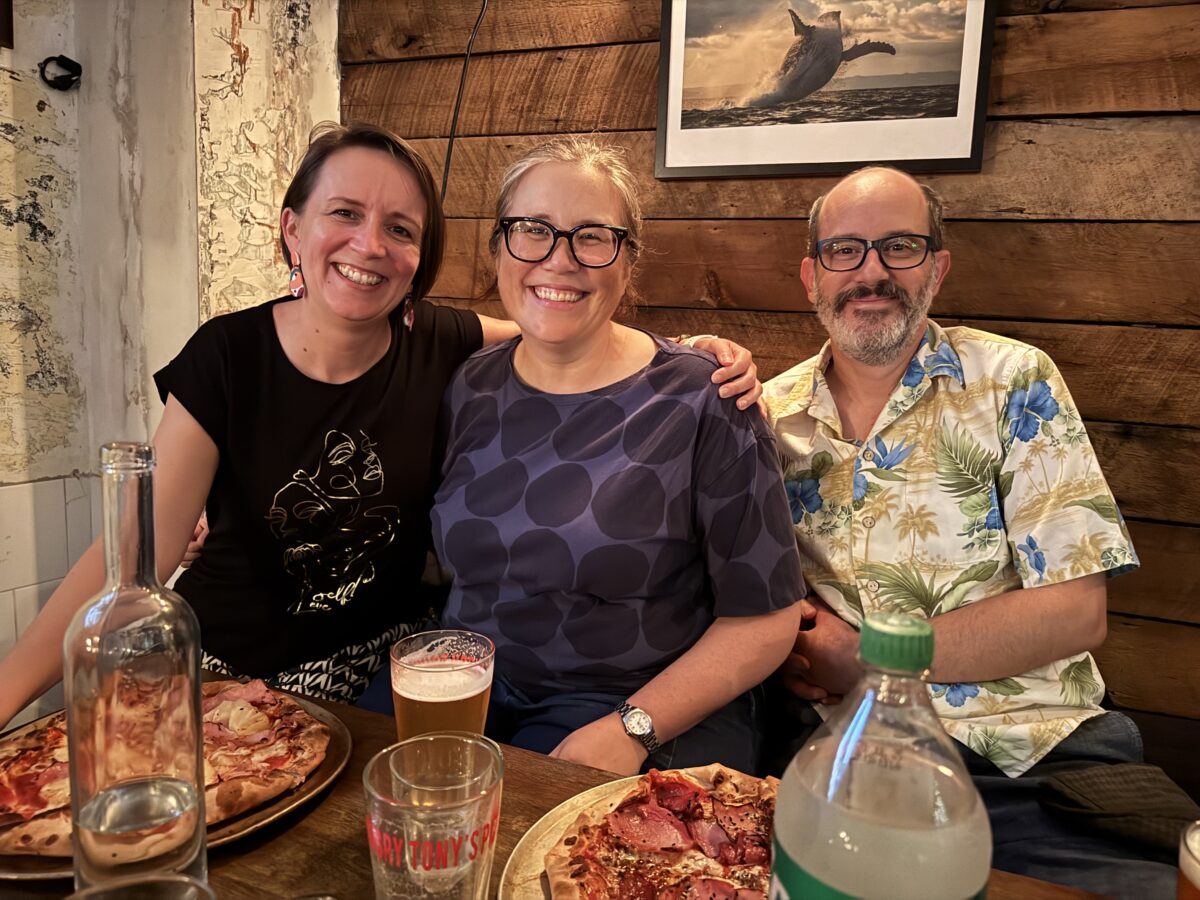 Three people in a restaurant, with pizza and drinks on the table in front of them