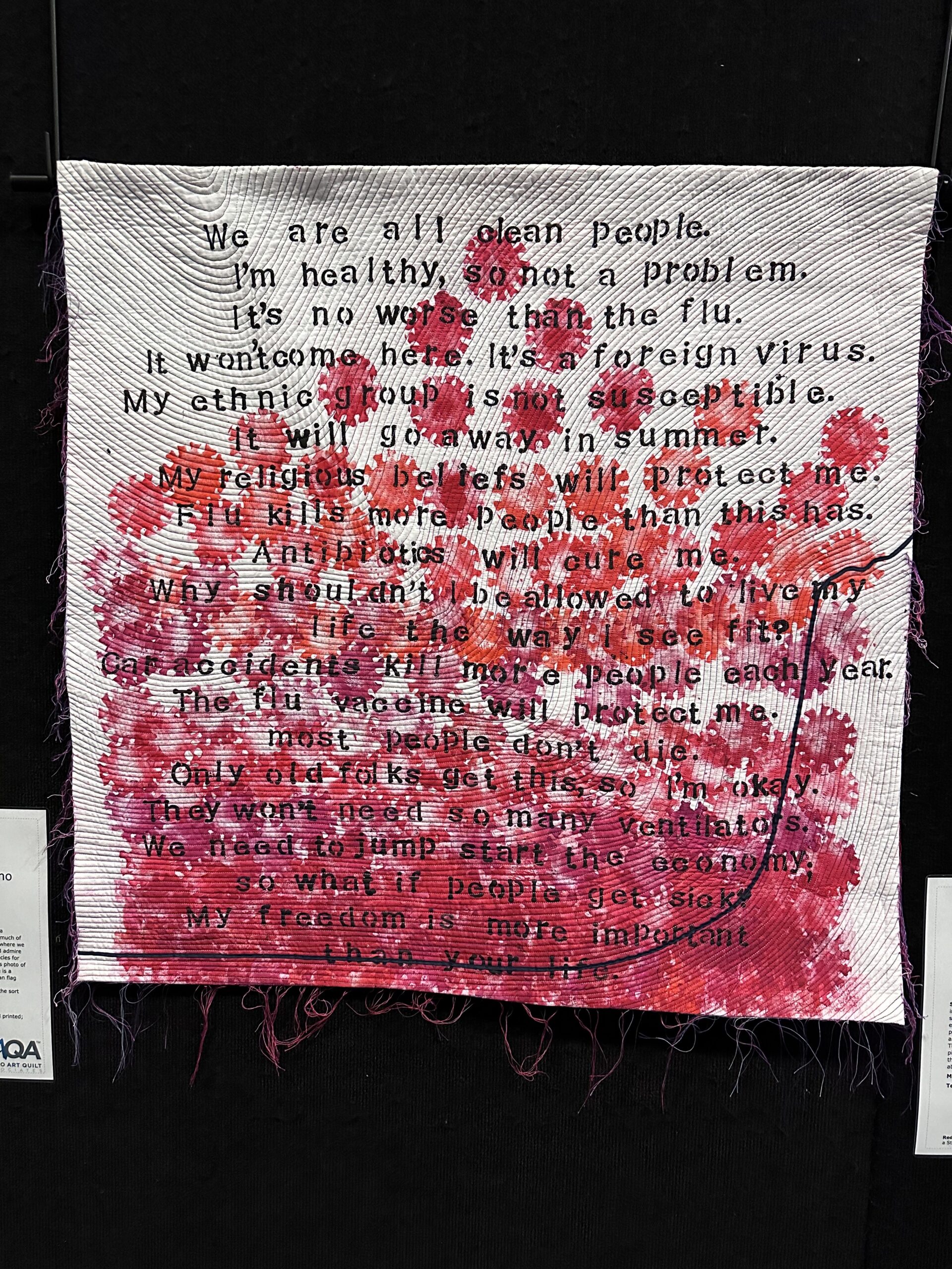 A quilt referencing the Covid pandemic with red flowers over a litany of excuses for not masking 