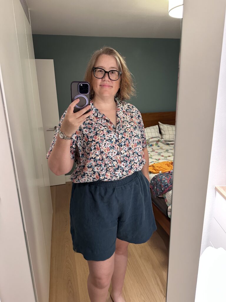 A selfie of a woman in blue linen shorts with a tucked-in printed button-down short-sleeved shirt