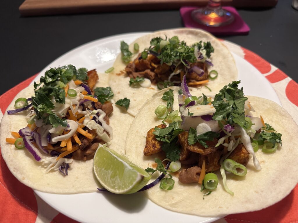 A plate with three flour tortillas piled with refried beans, roasted sweet potatoes, slaw, and coriander