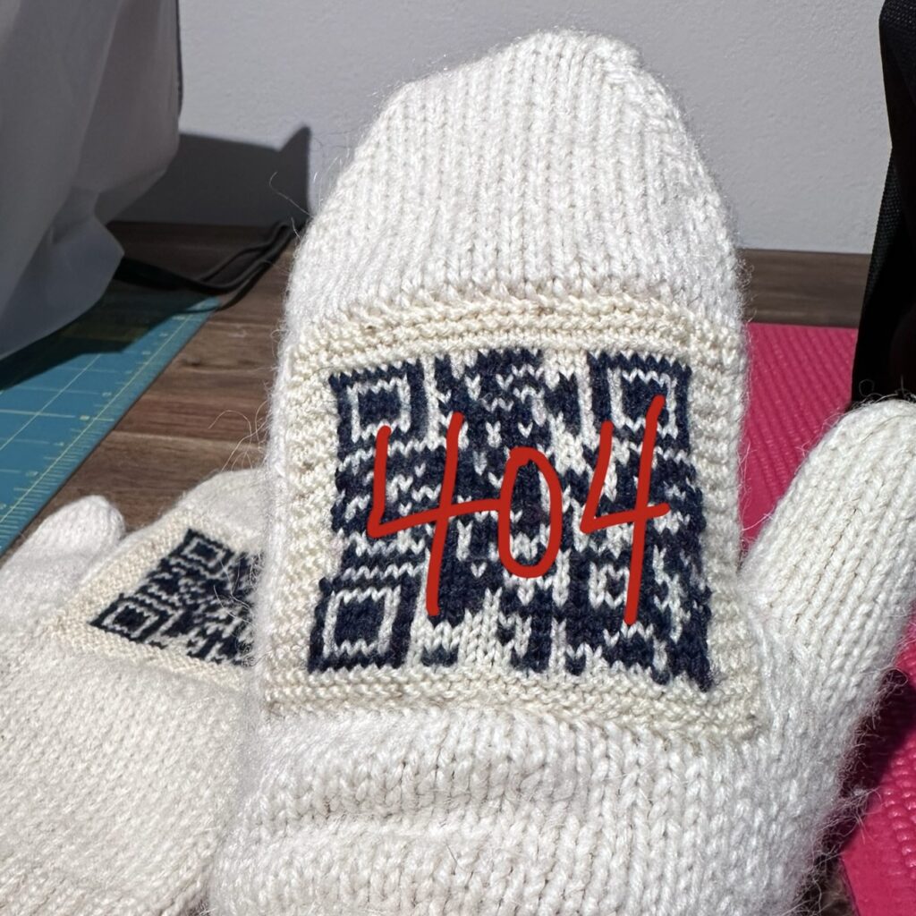 A knitted pair of mittens with a QR code. "404" is written over the image in red marker.