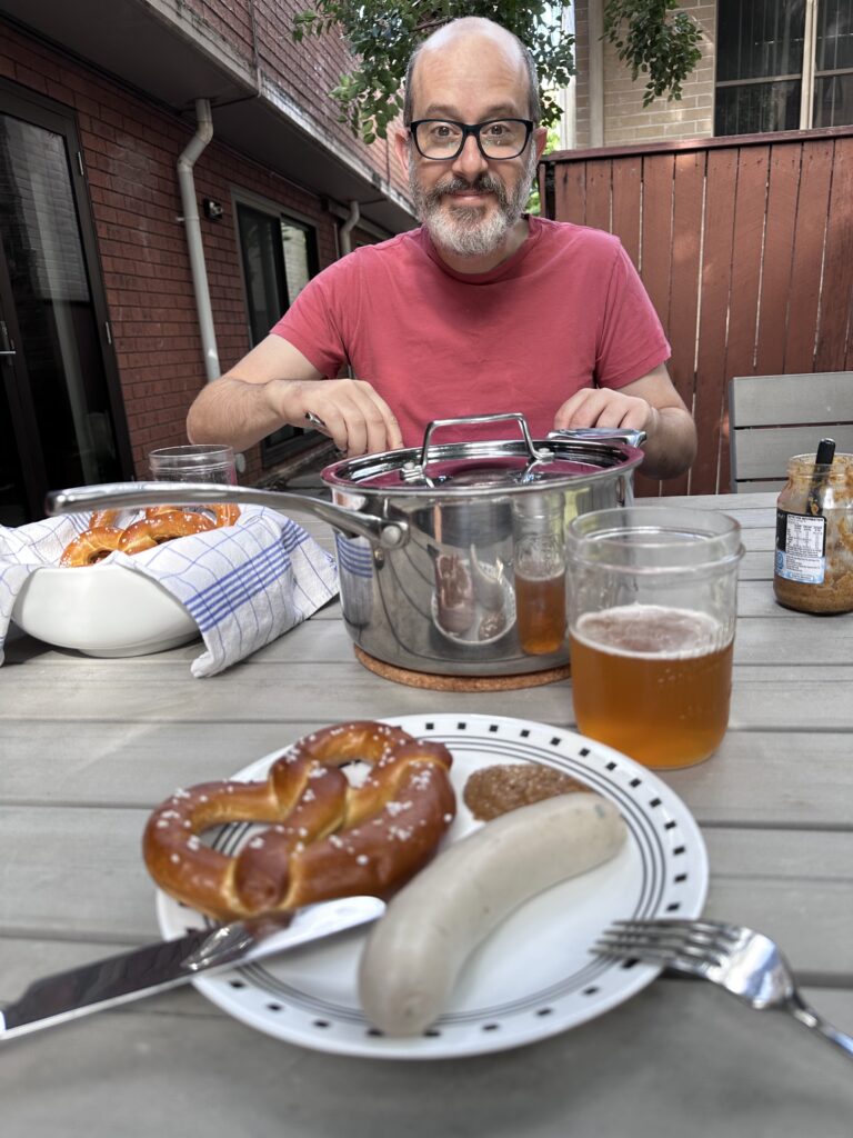 A man sits at a table outside. In front of him is a stainless steel lidded pot, a bowl of baked Bavarian pretzels, a jar of mustard, beers, and the plate of his companion.