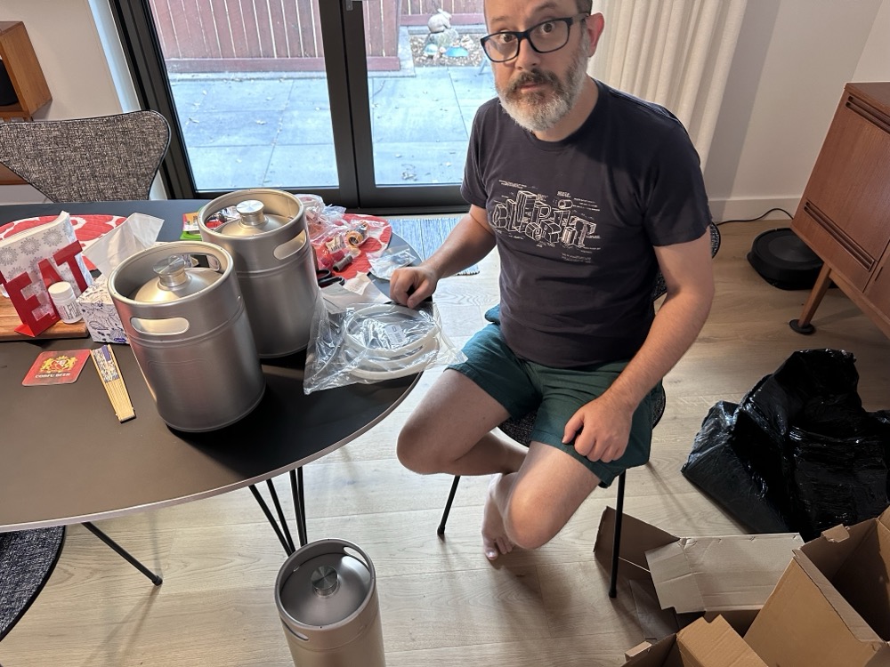 A beardy man sitting at a table with some shiny metal mini beer kegs