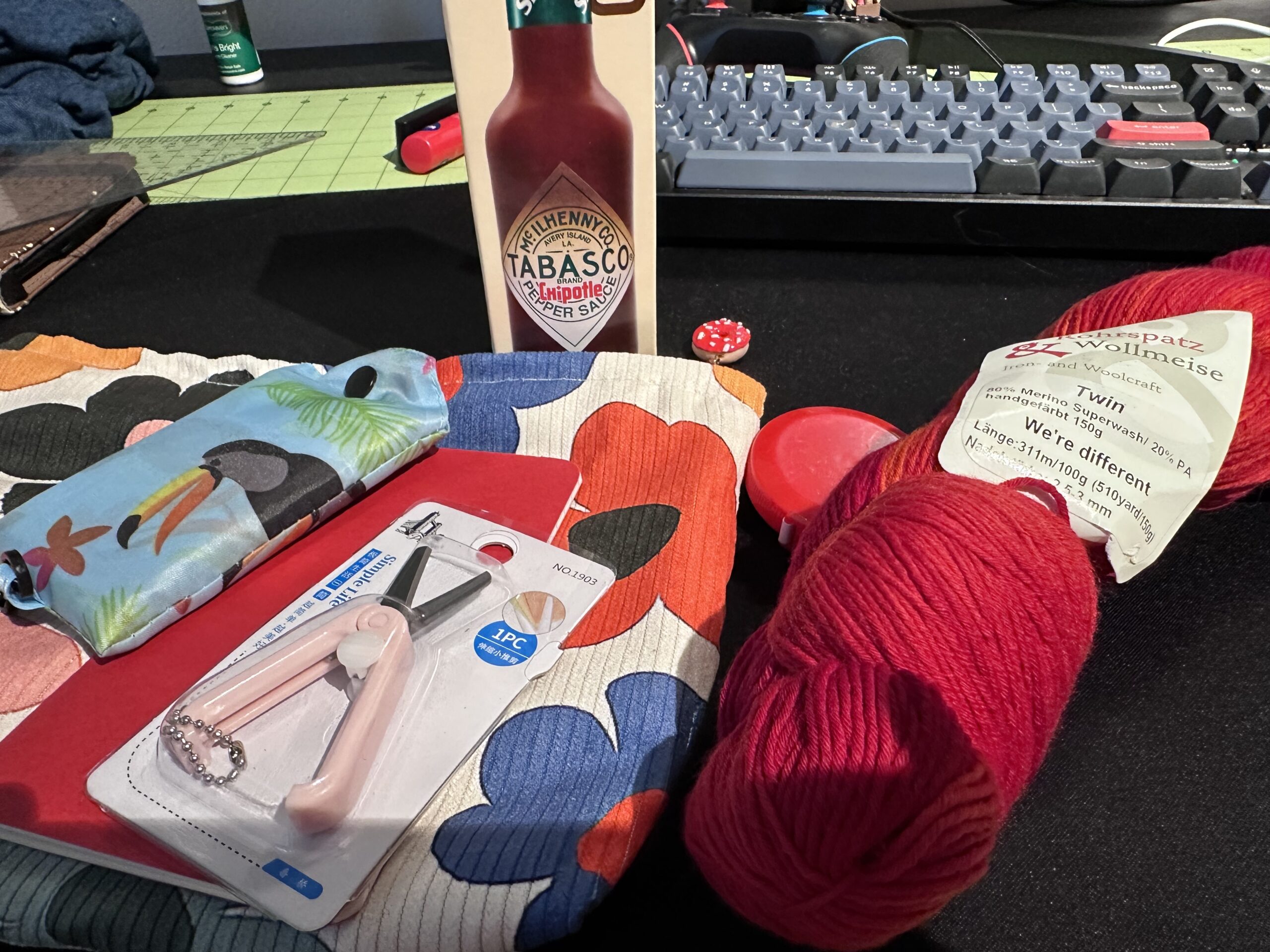 A skein of red Wollmeise knitting yarn, a large bottle of Tabasco Chipotle Hot sauce, and a pile of knitting things