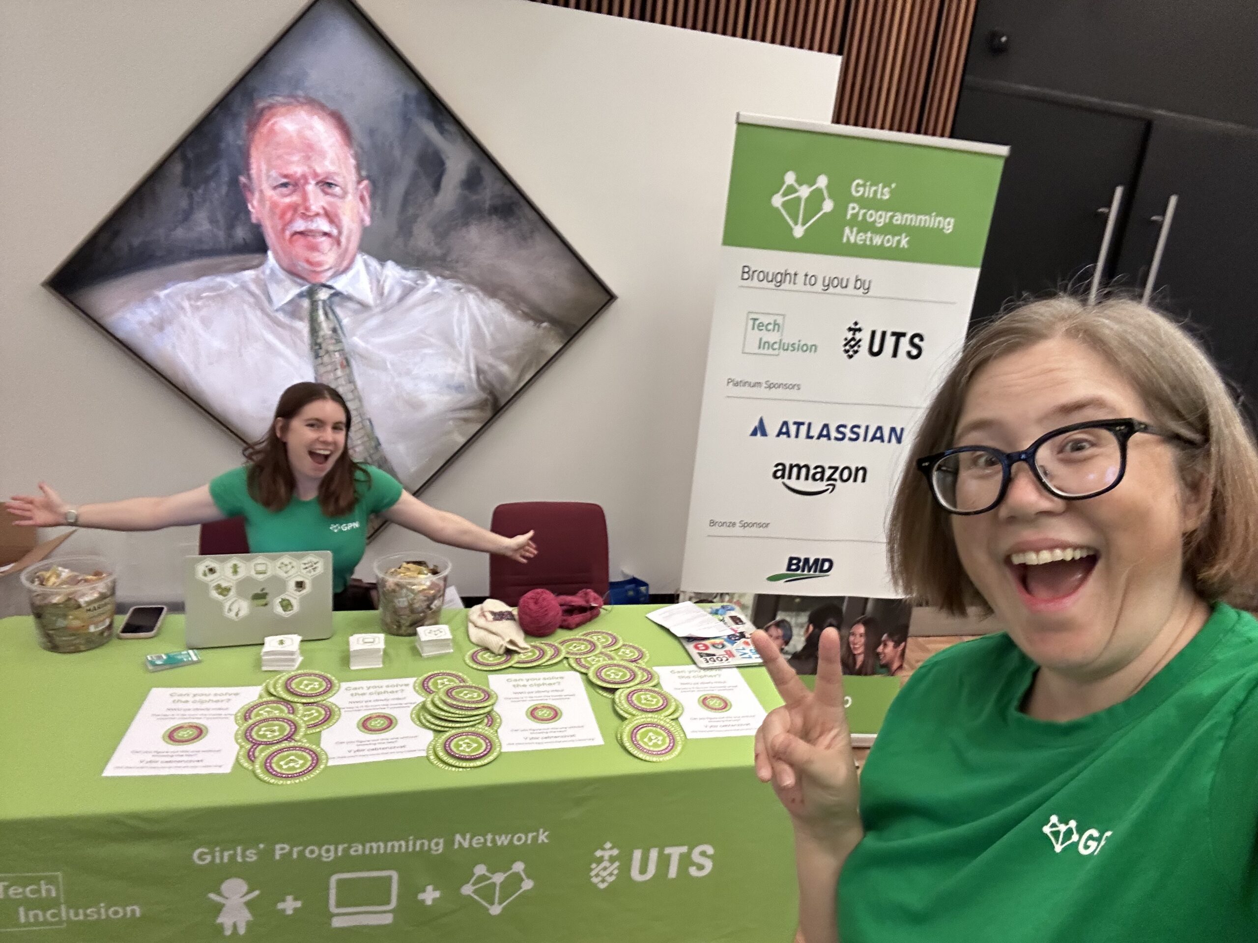 Two women in green shirts at a green conference booth. There’s also a weird painting of a dude behind them.