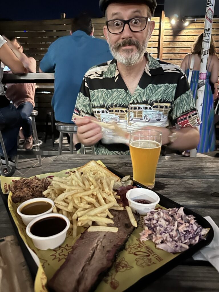 A man in an Aloha shirt with a tray full of bbq in front of him: brisket, pork, coleslaw, fries, pickles, and bread